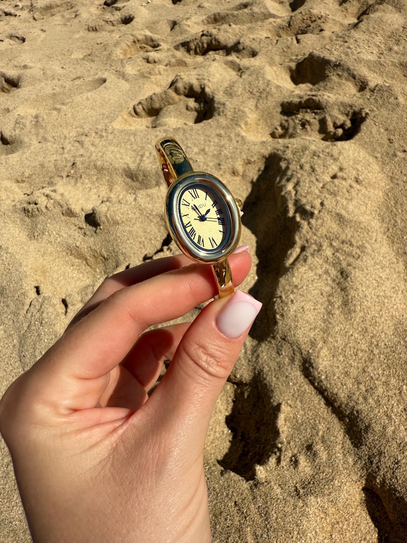 May include: Gold-toned oval wristwatch with a cream-colored face and Roman numeral hour markers. The watch has a gold band and is held against a sandy background. The watch face displays the time.