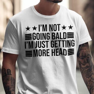May include: White t-shirt with black text that reads "I'm not going bald I'm just getting more head" with three stars and three stripes on each side of the text.