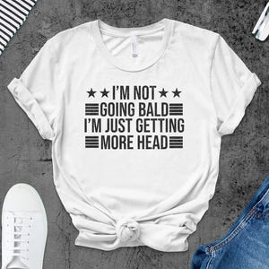 May include: White t-shirt with black text that reads "I'm not going bald I'm just getting more head" with three stars above and below the text.