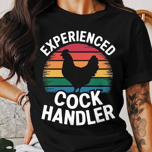 May include: Black t-shirt with a graphic featuring a rooster silhouette against a sunset design in red, orange, yellow, green, and blue. The text reads "EXPERIENCED COCK HANDLER" in white.