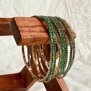 May include: A display of gold and green bangles on a wooden stand. The bangles are arranged in stacks, with some featuring gold links and others adorned with green gemstones. The wooden stand has a natural finish.