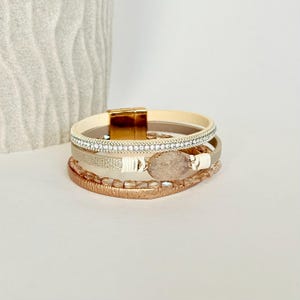 May include: A multi-strand bracelet featuring a cream-colored leather band with a gold magnetic clasp. The bracelet includes a strand with small rhinestones, a textured band, a band with a light-colored stone, and a copper-colored beaded strand.