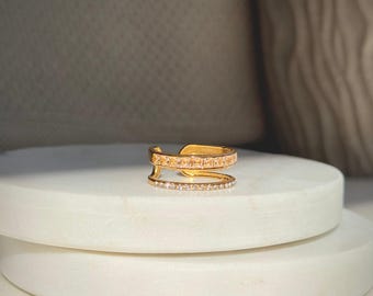 May include: A gold-colored ring with a double-band design. The top band features a row of square-cut, clear stones, while the lower band is adorned with smaller, round, clear stones. The ring is displayed on a white surface.