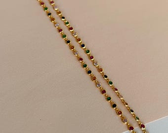 May include: Two delicate gold necklaces with colorful gemstones. The gemstones are in a variety of colors, including red, green, blue, and clear. The necklaces are displayed against a light pink background.