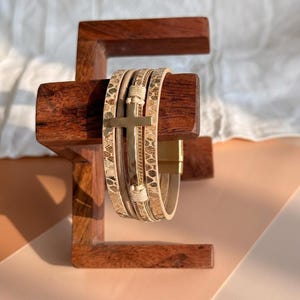 May include: A multi-strand wrap bracelet featuring a gold-toned metal cross charm centered on a band with a tan and brown snakeskin-like pattern. The bracelet includes thin gold chains and light-colored cord accents with a magnetic clasp closure.