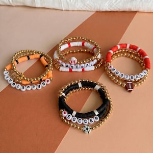 May include: Four sets of beaded bracelets featuring gold-tone metallic beads and colorful clay discs. Each set includes letter beads spelling BASKETBALL, BASEBALL, FOOTBALL, or SOCCER, accompanied by small charms shaped like the respective sports balls.