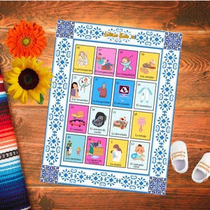 Op de afbeelding: Een kleurrijk Loteria-spelbord met verschillende illustraties en Spaanse tekst, met een blauw-witte decoratieve rand. Het bord staat op een houten oppervlak, met bloemen en babyschoentjes in de buurt.
