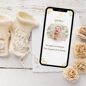 May include: A smartphone displays a baby's photo with the name "Mateo" and Spanish text. Cream-colored knitted baby booties with floral and pearl accents are next to the phone. Decorative beige paper flowers are also present.