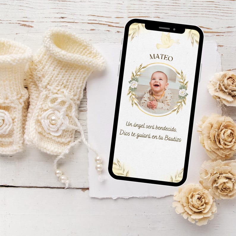 May include: A smartphone displays a baby's photo with the name "Mateo" and Spanish text. Cream-colored knitted baby booties with floral and pearl accents are next to the phone. Decorative beige paper flowers are also present.