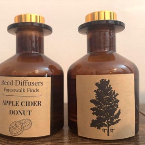 May include: Two amber glass reed diffuser bottles with gold-colored caps. The left bottle features a tan label reading Reed Diffusers Forestwalk Finds APPLE CIDER DONUT with a donut illustration. The right bottle has a tan label with a black tree graphic.
