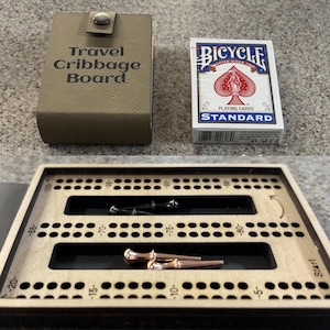 Könnte beinhalten: Ein beiges Reise-Cribbage-Board-Etui mit Druckknopfverschluss, ein Kartenspiel von Bicycle Standard und ein offenes Cribbage-Board aus Holz mit nummerierten Bahnen, zwei silberfarbenen Stiften und zwei kupferfarbenen Stiften.