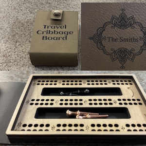 Könnte beinhalten: Ein klappbares Reise-Cribbage-Brett mit beigefarbener Lederoptik und der Aufschrift Travel Cribbage Board sowie The Smiths. Das Innere besteht aus hellem Holz mit nummerierten Bahnen und enthält zwei schwarze und zwei kupferfarbene Metallstifte.