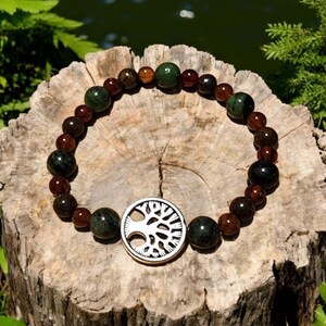 May include: A beaded bracelet with brown and green stones and a silver tree of life charm. The bracelet is on a wooden surface.