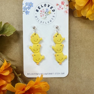 May include: A pair of yellow chick-shaped earrings with silver posts. Each earring has three stacked chicks with orange beaks and feet. The earrings are on a white card with the text "BELOVED WILDFLOWER" and the Etsy shop URL.