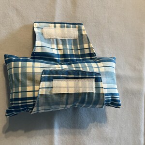 May include: A rectangular fabric pouch featuring a blue and white plaid pattern. The pouch includes a fold-over flap secured with a white hook-and-loop fastener strip, and a matching front pocket also secured with a white hook-and-loop fastener strip.