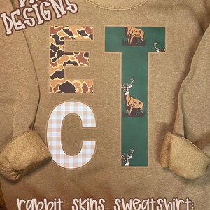 Puede incluir: Sudadera marr&oacute;n coyote con las letras "ETC". La "E" tiene un patr&oacute;n de camuflaje, la "T" un gr&aacute;fico de ciervo y la "C" un dise&ntilde;o de cuadros. Tambi&eacute;n se ve el texto "DKR DESIGNS" y "rabbit skins sweatshirt: coyote brown".