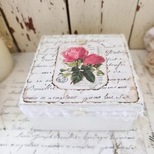May include: A square white wooden box featuring a decoupage design of pink roses on the lid. The lid is decorated with vintage-style cursive script text and a white lace trim along the edge. A small white button is attached to the top center.