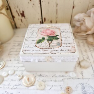 May include: A small, square, off-white wooden box featuring a central rectangular label with a pink rose illustration and cursive script text. The lid is decorated with a white lace trim and a single button centered on the front edge.