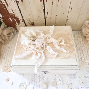 May include: A rectangular off-white wooden box featuring a decorative top panel adorned with a cream-colored fabric flower, a single large pearl-like bead, a strand of small white faux pearls, and a strip of beige lace with trailing fabric ribbons.