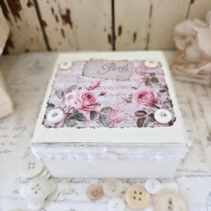 May include: A square off-white wooden box featuring a decorative top panel with pink roses and the text Paris. The lid is accented with four white buttons at the corners and a white lace trim along the edge. The box has a distressed paint finish.