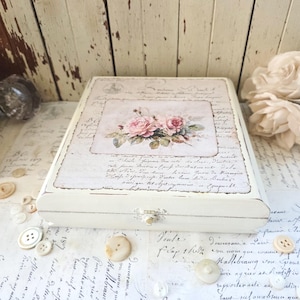 May include: A rectangular white wooden box featuring a central illustration of pink roses surrounded by vintage-style cursive French script text. The box has a small metal latch on the front and is surrounded by several loose white buttons.