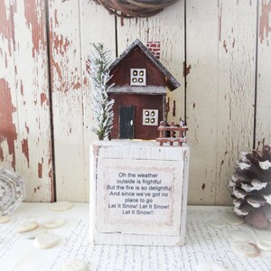 May include: A miniature wooden house with a red roof, chimney, and a small fence. The house sits atop a white block with the text "Oh the weather outside is frightful...Let It Snow!" A small evergreen tree is to the left of the house.