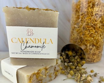 May include: Two bars of handmade soap with the words "CALENDULA Chamomile" on the label. The soap is light beige with dried flower petals on top. A glass jar filled with dried flowers and a small metal scoop with dried chamomile flowers are in the background.
