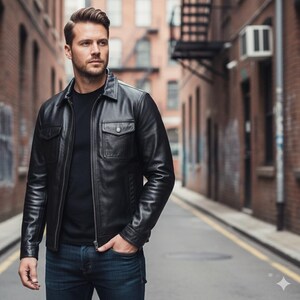 May include: Black leather jacket with two chest pockets and a zipper closure. The jacket is worn with a black shirt and blue jeans. The person is standing in an alley with brick buildings.