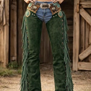 May include: Green suede chaps with long fringe detail. The chaps are worn with a decorative belt with turquoise accents and a silver buckle. The model is wearing blue jeans and cowboy boots.