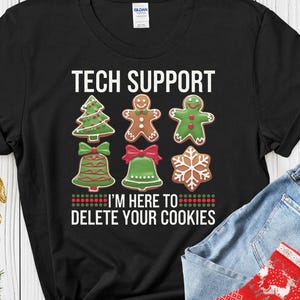 Może przedstawiać: Czarny t-shirt z napisem "TECH SUPPORT" i "I'M HERE TO DELETE YOUR COOKIES". Koszulka przedstawia wzory piernikowych ciasteczek w kształcie choinki, ludzika z piernika, gwiazdki, dzwonka i płatka śniegu. Widoczne są również niebieskie dżinsy i czerwona świąteczna skarpetka.