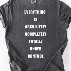 May include: Dark gray t-shirt with rolled-up sleeves. The shirt features the white text: "EVERYTHING IS ABSOLUTELY COMPLETELY TOTALLY UNDER CONTROL". The shirt is made of a soft, comfortable material.