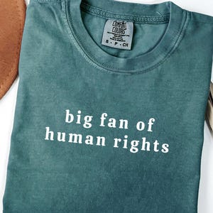 Pode incluir: T-shirt Comfort Colors verde-azulado dobrada com o texto branco "big fan of human rights". Um chapéu castanho e folhas secas são parcialmente visíveis. Etiqueta pequena perto da gola.