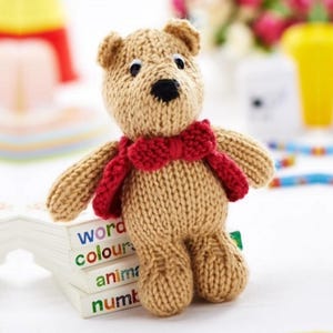 May include: A tan knitted teddy bear toy featuring a bright red bow tie around its neck. The bear has black plastic eyes and a black stitched nose. It is standing on a stack of four white books with the words word, colours, anima, and numb.