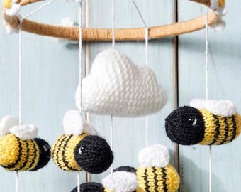May include: A hanging mobile featuring a circular frame wrapped in tan yarn, decorated with white and yellow crocheted flowers. Suspended from the frame are several crocheted bees with yellow and black stripes and white wings, along with a central white crocheted cloud.