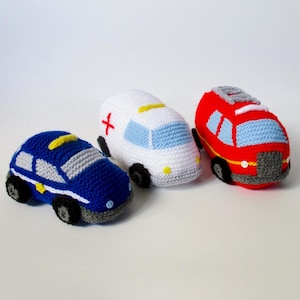 May include: Three crocheted toy vehicles including a blue police car, a white ambulance with a red cross, and a red fire engine. Each vehicle features stitched details, small button accents for lights or wheels, and grey yarn bases.