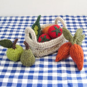 May include: A collection of handmade knitted play food items including a light green apple, a dark green pear, and two orange carrots with green tops. A beige crocheted basket contains a red tomato, a green pea pod, and an orange vegetable.