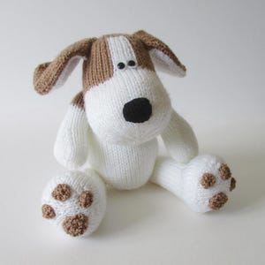May include: A knitted stuffed dog toy featuring a white body and head with brown patches on the ears and top of the head. The toy has a black nose, two small black eyes, and white paws with brown circular knit details representing paw pads.