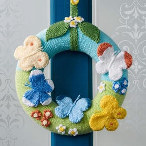May include: A circular knitted wreath featuring a light blue and lime green base decorated with various three-dimensional butterflies in yellow, white, orange, and blue. Small white and yellow flowers and green leaves are attached to the surface.