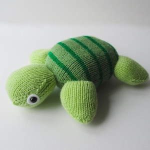 May include: A knitted turtle plush toy featuring a light green head and limbs with a darker green striped shell. The turtle has a single visible black and white plastic safety eye on the side of its head, crafted from soft yarn material.