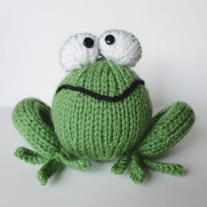 May include: A hand-knitted green frog toy featuring a round body, two large white eyes with black button pupils, and a stitched black mouth. The frog has four splayed, knitted green legs extending from the sides of its spherical, textured body.