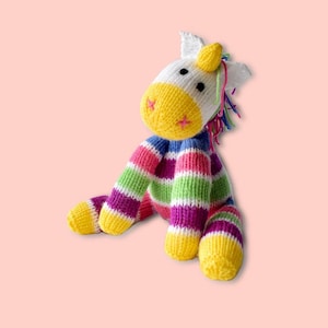 May include: A colourful striped knitted unicorn toy with a yellow face, white body, and rainbow mane. The unicorn is sitting with its legs crossed.