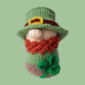 May include: A green knitted leprechaun doll wearing a green hat with a gold buckle and a red band. The doll has a red beard and is holding a green shamrock.
