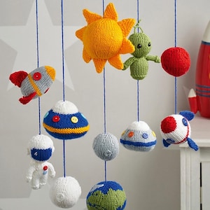 May include: A space-themed mobile featuring knitted figures of a yellow sun, green alien, red planet, rockets, UFOs, an astronaut, and a globe suspended from a blue circular frame by blue strings. The figures are crafted with yarn and buttons.