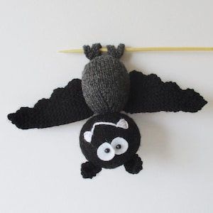 May include: A knitted bat toy with a dark grey body, black wings, and a black head. The bat features large white circular eyes with black pupils and two small white embroidered fangs. It is suspended from a thin, light-coloured wooden stick.