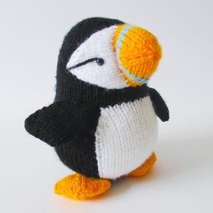 May include: A knitted puffin toy featuring a black back and wings, a white chest and face, and a bright orange beak with a light blue stripe. The bird has a small black eye and stands on two orange webbed feet, crafted from soft yarn.