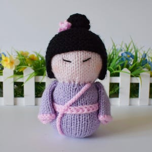 May include: A hand-knitted doll featuring a pale pink face with closed eyes, black hair styled in a bun with a small pink flower accessory, and a light purple outfit with a pink sash tied around the waist and pink cuffs on the sleeves.