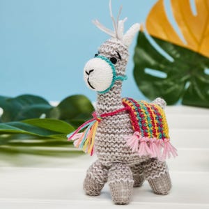 May include: A hand-knitted gray llama figurine featuring a white muzzle, a teal halter, and a colorful striped saddle blanket with pink fringe tassels. The llama has upright ears and a textured yarn construction, standing on four short legs.