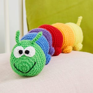 May include: A multi-coloured crochet caterpillar toy featuring a green head with white and black eyes and a stitched smile. The body consists of segments in blue, purple, red, orange, and yellow, each with a rounded shape and a small tail at the end.