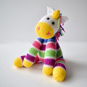 May include: A knitted unicorn toy featuring a white head with a yellow muzzle, pink stitched nostrils, and a yellow horn. The body and limbs are patterned with horizontal stripes in yellow, green, pink, blue, and purple, with a multi-coloured yarn mane.