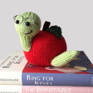 May include: A handmade knitted apple-shaped plush toy with a green worm wearing round glasses. The apple is red with a brown stem and green leaf. The toy is resting on top of two books with the title "Ring for Jeeves".